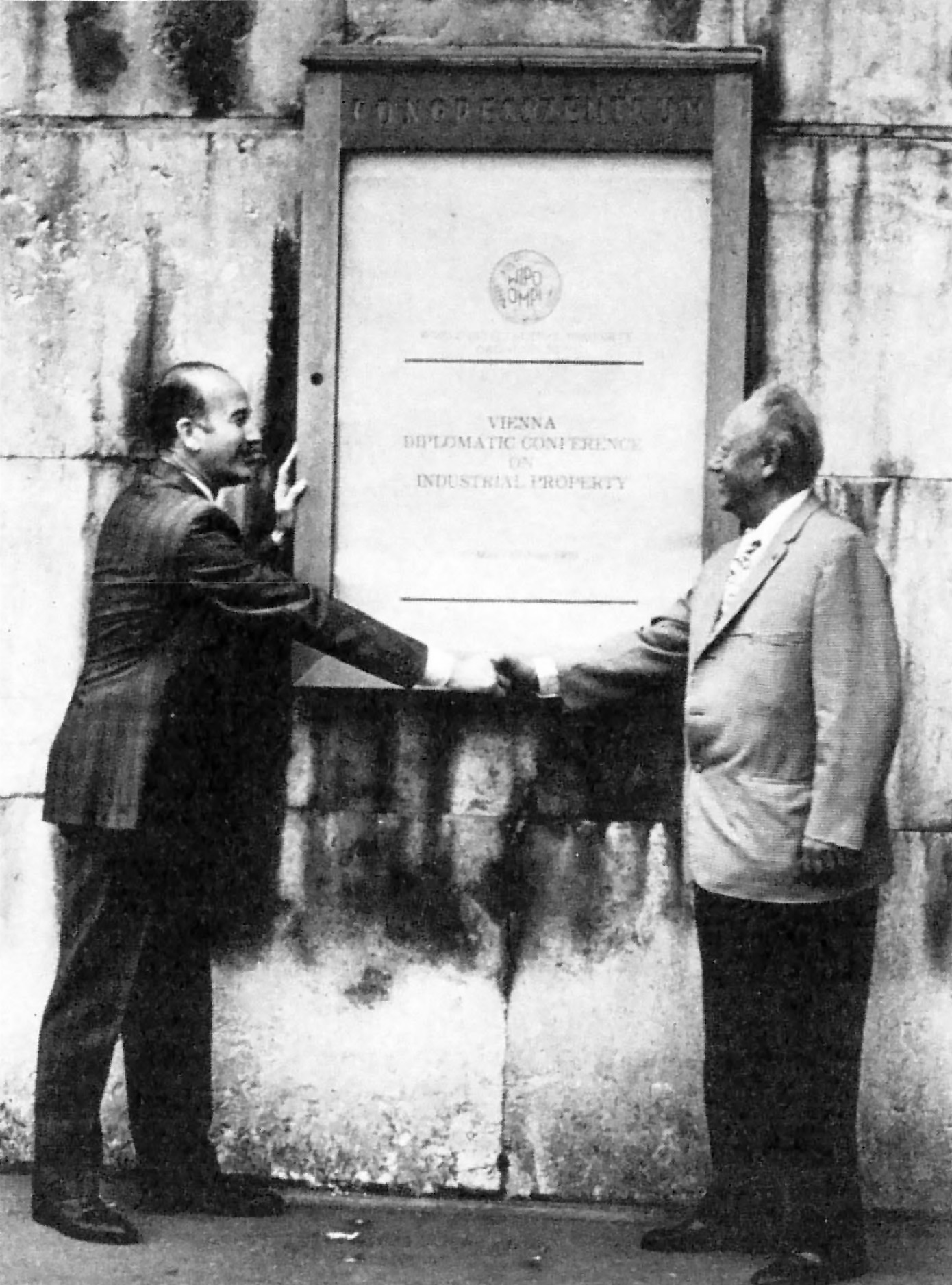 Black-and-white photograph of John Dreyfus (left) shaking hands with Charles Peignot (right) under a large wall plaque displaying a sign that says VIENNA DIPLOMATIC CONFERENCE ON INDUSTRIAL PROPERTY.